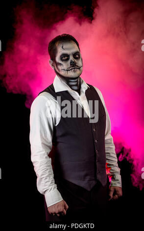 Porträt von einem gut gekleideten Mann mit Totenkopf Make-up. Horror und Halloween Motto, mit Bühnenbeleuchtung und Effekte. Farbige Nebel hinter dem Mod. Stockfoto