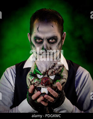 Porträt von einem gut gekleideten Mann mit Totenkopf Make-up Halten des Gesicht von einem bösen Clown. Horror und Halloween Motto, mit Bühnenbeleuchtung und e Stockfoto