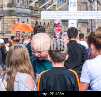 Edinburgh, Schottland, Großbritannien. 9. August 2018. Edinburgh Fringe Festival, Royal Mile, Edinburgh, Schottland, Vereinigtes Königreich. An einem sonnigen Festival Tag der Jungfrau Geld gesponsert Street Festival mit Menschen und fringe Darsteller verpackt ist. Eine Franse Interpret tragen eine grausame baby Kopf interagiert mit Fußgängern verteilen Flyer Stockfoto