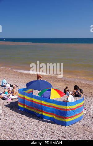 Urlauber auf Sidmouth Strand mit Wind brechen und Sonnenschirme. Auf der Suche über eine Trübe im Englischen Kanal. East Devon, Großbritannien. August, 2018. Stockfoto