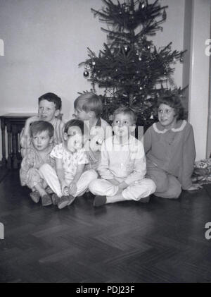 1960 s, neugierige junge chiildren auf einen Holzboden, der ein Zimmer an Weihnachten in Ihren Schlafanzug und Hausschuhen vor einem geschmückten Weihnachtsbaum Sitzen, Warten auf ihre Geschenke, England, UK. Stockfoto