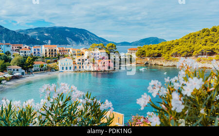 Panoramablick nach Assos Dorf in Kefalonia, Griechenland. Helle, weiße Blüte Blume im Vordergrund der türkisfarbenen ruhige Bucht von Mittelmeer und schöne bunte Häuser im Hintergrund Stockfoto