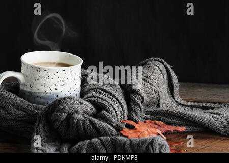 Heiß dampfende Tasse Kaffee mit Sahne im gemütlichen grau Schal mit Laub im Herbst gewickelt. Extrem flache Tiefenschärfe mit selektiven Fokus