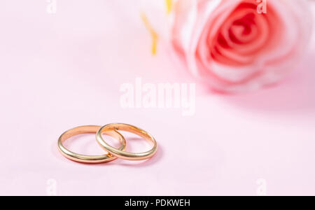 Liebe und Ehe Konzept. Nahaufnahme der Goldenen Hochzeit Ringe und eine verschwommene rosa Rose, kopieren, auf einem rosa Hintergrund. Stockfoto