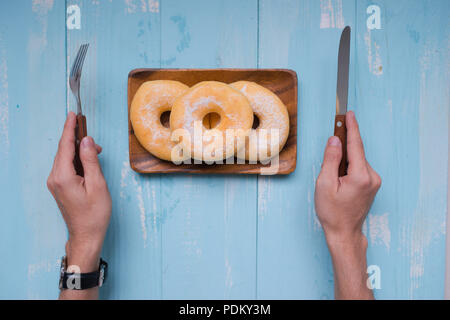 Frühstück Konzept: Nahaufnahme der männlichen Händen mit Gabel und Messer schneiden Donut Form Keks auf Blau Holz- Hintergrund; Ansicht von oben, flach Stockfoto