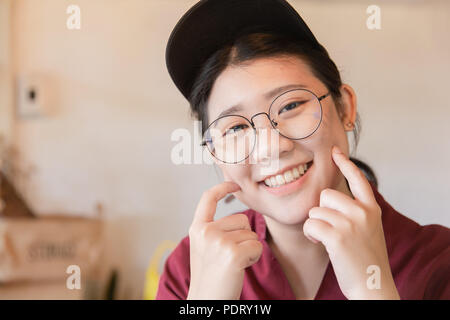 Chubby plump jugendlich niedlichen weißen Zähnen lächeln Asiatische junge Studentin mit Brille und Hut finger Wange mit Kopie Raum Stockfoto