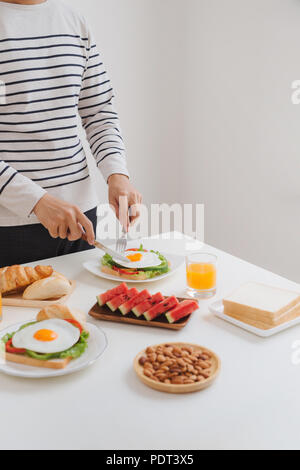 Die Hände des Menschen schneiden Sie die Eier auf Papier, serviert mit Brot, Kräutern, Wurst und frischem Saft. Stockfoto