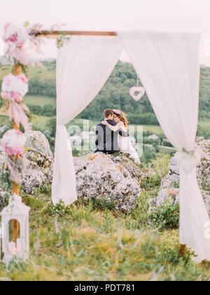 Die Rückansicht der fröhlichen Brautpaar sitzt auf dem grossen Felsen an der Vorderseite des Bogens. Stockfoto