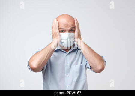 Portrait von reifer Mann tragen medizinische Gesundheit Maske, auf weißem Hintergrund. Er schließt seine Ohren mit den Händen. Schockiert Mann emotion Stockfoto