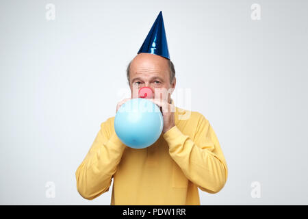 Portrait von reifer Mann im gelben T-Shirt und lustige Kappe weht eine blaue Ballon über einen weißen Hintergrund. Die Vorbereitung für eine Geburtstagsfeier Stockfoto