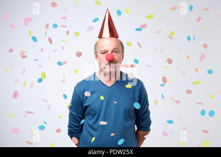 Unglücklich Geburtstag älterer Mann ist traurig und enttäuscht, weil niemand kam, um seinen Geburtstag zu feiern. Blasen party Horn, lustige Kappe und rote Clownnase Stockfoto