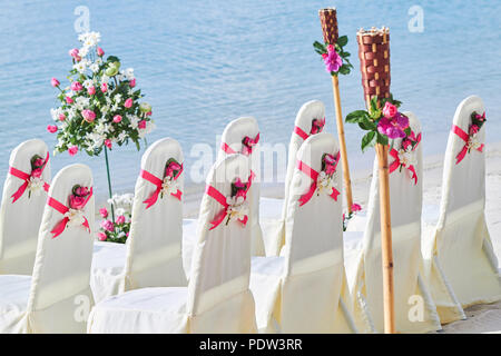 Wieder eine Gruppe von Weißen Hochzeit Stühle Seite schmücken mit rot rosa Organza Schärpe mit Konus von Rosen Blütenblatt, Strand Hochzeit Einstellungen Stockfoto