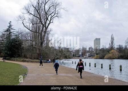 Zwei Kinder laufen auf dem Weg in die Kensington Gardens, neben dem See Serpentine Lake, im Winter. Andere Menschen zu Fuß und Paar sitzt auf der Bank. Stockfoto