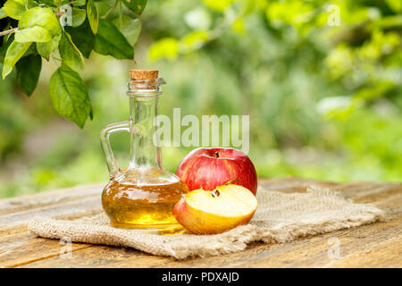 Apfelessig in Glasflasche mit Korken und Frische rote Äpfel am Sack und alte hölzerne Bretter mit Apfelbäumen im Hintergrund. Bio-lebensmittel für h Stockfoto