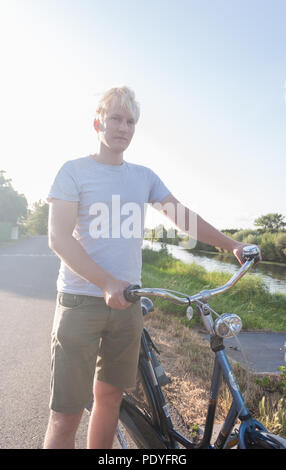 Ein junger Mann steht mit seinem Fahrrad in der Nähe eines Flusses an einem späten Sommernachmittag. Stockfoto