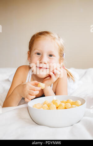 Porträt einer weiße Kaukasier Kind Mädchen lächelnd Essen Mais Puffs. Preschooler kid essen Snacks fast food im Bett zu Hause im Innenbereich. Gesunde Mahlzeit Chil Stockfoto