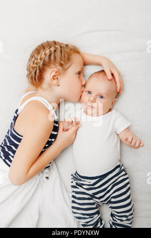 Lifestyle Portrait von niedlichen weißen Kaukasische Mädchen Schwester Holding küssen kleine Baby, liegend auf dem Bett drinnen. Ältere Geschwister mit jüngeren Bruder Neugeborenen. Stockfoto