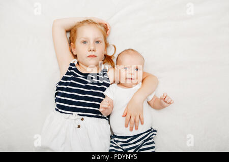 Lifestyle Portrait von niedlichen weißen Kaukasische Mädchen Schwester Holding Baby, liegend auf dem Bett. Ältere Geschwister mit jüngeren Bruder Schwester neugeborenen Suchen i Stockfoto