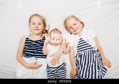 Lifestyle Portrait von niedlichen weißen Kaukasische Mädchen Schwestern halten kleines Baby, liegend auf Bett Decke im Innenbereich. Ältere Geschwister mit jüngeren Bruder Schwester Stockfoto