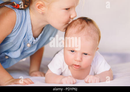 Lifestyle Closeup Portrait von niedlichen weißen Kaukasische Mädchen Schwester küssen kleine Baby, liegend auf dem Bett drinnen. Ältere Geschwister mit jüngeren Bruder Neugeborenen. Stockfoto
