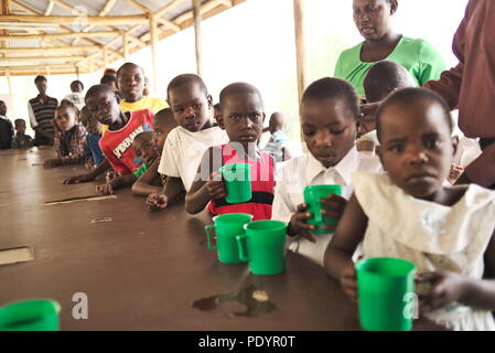 Junge afrikanische Kinder warten in einer Warteschlange vor einem ugandischen Krankenhaus auf Essen und Getränke mit Plastikbechern und Schalen, die miserabel aussehen Stockfoto