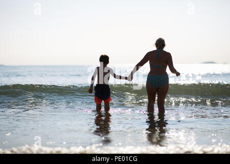 Mutter und Sohn Spaß springen die Wellen des Meeres in den frühen Abend, als die Sonne untergeht und glitzert auf die reinen blauen Wasser Stockfoto