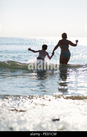 Mutter und Sohn Spaß springen die Wellen des Meeres in den frühen Abend, als die Sonne untergeht und glitzert auf die reinen blauen Wasser Stockfoto