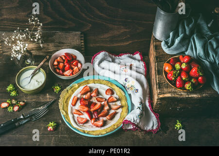 Hausgemachte Crepes auf dunklen rustikalen Küche Tisch mit Erdbeeren und Joghurt auf blauen Platte. Country Style food Still Life Stockfoto