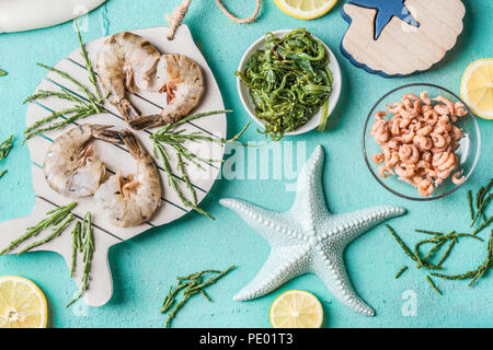 Tiger Garnelen und Nordsee Krabben auf hellblauem Hintergrund mit Algen in Schalen, Ansicht von oben, flach. Meeresfrüchte kochen Konzept Stockfoto