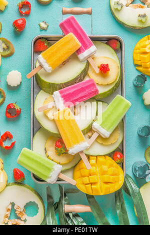 Fach mit bunten Eis Eis am Stiel mit frisch geschnittenem Obst und Beeren Zutaten auf hellblauem Hintergrund, Ansicht von oben, flach. Gefrorenen tropischen Stockfoto
