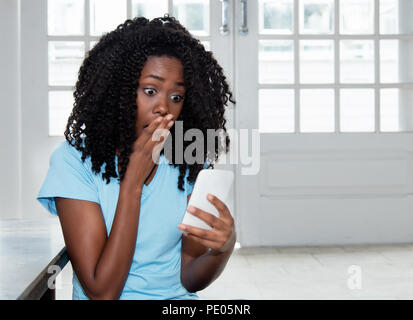 Afrikanische amerikanische Frau, die schlechten Nachrichten auf dem Telefon zu Hause schockiert in Innenräumen Stockfoto