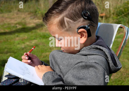 Seitenprofilansicht eines jungen Kindes 8 mit coolem Sternhaarschnitt, das im Garten sitzt und ein Skizzenbuch zeichnet, das Cochlear-Implantate-Gerät UK KATHY DEWITT trägt Stockfoto