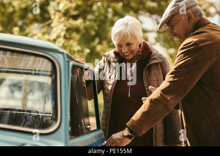 Interessieren gentleman tragen warmen Mantel und Hut öffnung Auto für seine Frau, während die zusammen reisen. Senior Paar gehen auf einen Road Trip. Stockfoto