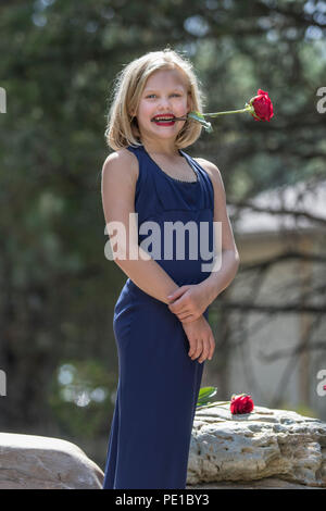 Hübsche, junge blonde Frau. preteenn, Mode schießen. Standing Rock, mit einer roten Rose in den Zähnen. 3/4 Profil, von der Kamera suchen, blaues Kleid, Modell Stockfoto