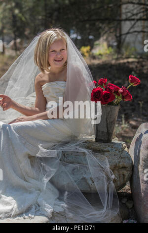 Fantasy, 8-9 Jahr attractie blond, mit einem Hochzeitskleid Kleid & Tante Schleier. Das Sitzen im Freien auf grossen Felsen neben Vase mit roten Rosen. Stockfoto