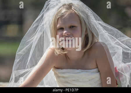 Fantasy, 8-9 Jahr attractie blond, mit einem Hochzeitskleid Kleid & Tante Schleier. Das Sitzen im Freien auf grossen Felsen neben Vase mit roten Rosen. Stockfoto