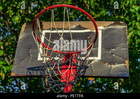 Alter und beschädigter Basketballkorb mit Käfig, Baum und Haus im Hintergrund, Ansicht von unten, in der Nähe Stockfoto