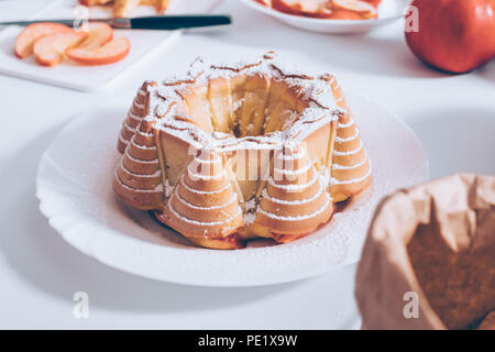 Close-up traditionelle Apfelkuchen mit Puderzucker über Zutaten und Utensilien auf weißen Tisch umgeben dekoriert. Hausgemachte Backwaren die Zusammensetzung von Lebensmitteln. Stockfoto