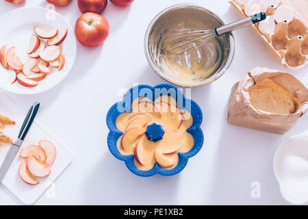 Auflaufform gefüllt mit Teig und Fruchtschnitten neben Metall Schüssel, Schneebesen, Eierschalen, Mehl und Zucker, flach. Zutaten und Werkzeuge für das Kochen ap Stockfoto