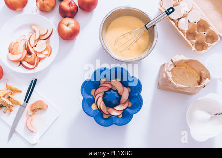 Silikon Form gefüllt mit Teig und Fruchtschnitten neben Metall Schüssel, Schneebesen, Eierschalen, Mehl und Zucker, flach. Zutaten und Werkzeuge für das Kochen Stockfoto