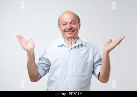 Indoor Porträt verwirrter älterer Mann im blauen Shirt, Ich habe keine Idee, Geste, zuckte mit den Schultern und Händen, stehend gegen Grau zurück Stockfoto