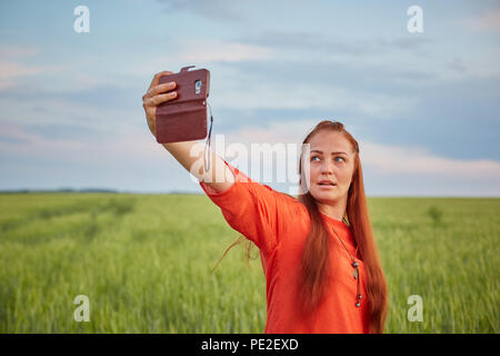 Junge schöne Frau im roten Kleid und rote Haare, nimmt eine selfie auf dem Handy in die Grüne Weizenfeld am Abend bei Sonnenuntergang im Sommer. Die Natur der digitalen Technologie. Stockfoto