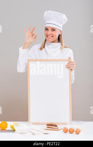 Schöne weibliche Konditor zeigt ok Zeichen halten Whiteboard mit Kuchen und Zutaten für den Kuchen auf den Tisch. Stockfoto
