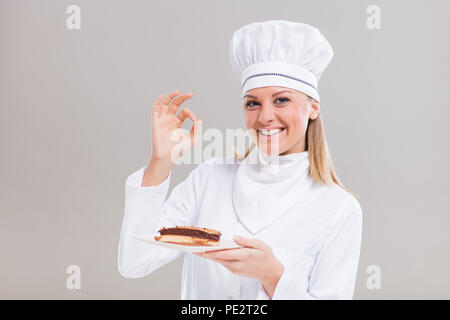 Schöne weibliche Konditor zeigt Stück Kuchen und ok Zeichen auf grauem Hintergrund. Stockfoto