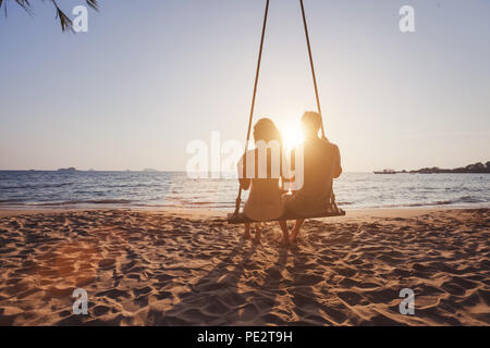 Urlaub am Meer für romantische junges Paar, Hochzeitsreise Urlaub, Silhouetten von Mann und Frau sitzen zusammen auf Swing und Sonnenuntergang Stockfoto