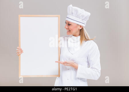 Porträt der schönen Köchin zeigt leeres Whiteboard auf grauem Hintergrund. Stockfoto