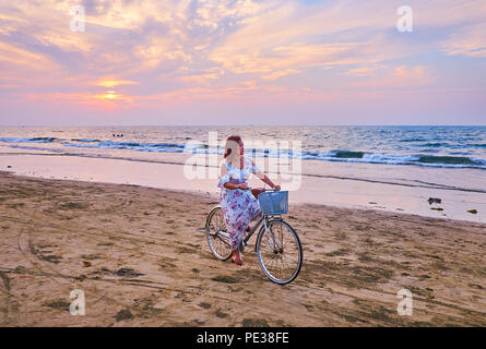 CHAUNG THA, MYANMAR - 28. FEBRUAR 2018: Die schöne junge asiatische Frau in langen, romantischen Kleid genießt den Zyklus Reiten am Strand entlang Linie auf Sonnenuntergang Stockfoto