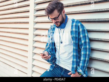 Trendy hipster Mann im blauen Hemd und Kopfhörer surfen Smartphone während lehnte sich an der Wand auf der Straße und lächelnd Stockfoto