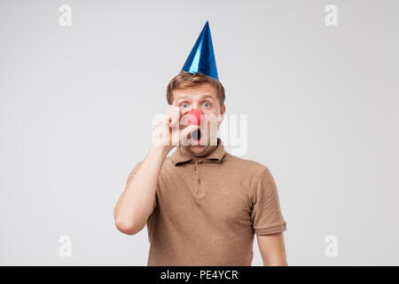 Kaukasischen jungen Mann mit Urlaub Kappe auf dem Kopf und roten clown Nase Grimassen und Spaß haben. Konzept der Geburtstagsfeier Stockfoto