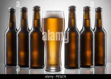 Glas und Flaschen Bier. Mit Tropfen, hohes Glas mit Sekt Bier und dicken Schaumstoff abgedeckt ist vor zwei Zeilen mit sechs braunen ungeöffnete Flaschen auf n Stockfoto